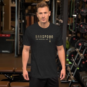 May include: A dark gray t-shirt with the gold text "BARSPOON" and "THE COCKTAIL APP" printed on the front. The shirt is short-sleeved and worn by a person in a gym setting.