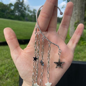 Może przedstawiać: Srebrny nausznik z wiszącymi łańcuszkami i zawieszkami w kształcie gwiazd. Nausznik owija się wokół ucha i posiada wiele łańcuszków o różnej długości. Zawieszki obejmują czarne i białe kształty gwiazd.