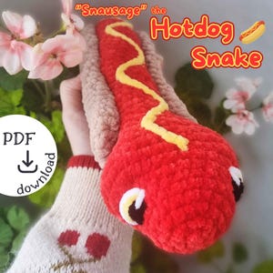 Peut inclure: Un jouet en peluche serpent hot-dog crocheté avec un corps rouge, de la moutarde jaune et un pain brun. Le jouet est tenu dans une main portant un pull avec un motif de cerise. Le texte "Snausage" the Hotdog Snake est écrit au-dessus du jouet.