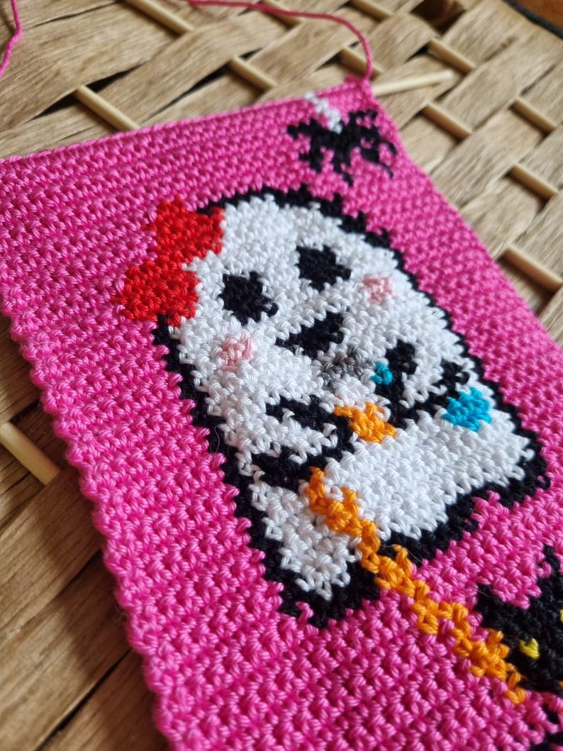 K&ouml;nnte beinhalten: Handgefertigtes, pinkfarbenes geh&auml;keltes Quadrat mit einem Cartoon-Geist-Design. Der Geist hat ein wei&szlig;es Gesicht mit schwarzen Augen und einem schwarzen Mund. Er tr&auml;gt eine rote Schleife und h&auml;lt einen blauen Gegenstand. &Uuml;ber dem Geist befindet sich eine schwarze Spinne.