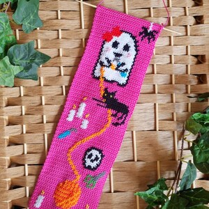 K&ouml;nnte beinhalten: Ein handgefertigter, pinkfarbener Lesezeichen zum Thema Halloween. Das Lesezeichen zeigt einen wei&szlig;en Geist mit roter Schleife, eine schwarze Katze, eine Spinne, Kerzen, einen Totenkopf und einen K&uuml;rbis. Das Lesezeichen ist aus H&auml;kelmaterial und hat eine pinkfarbene Aufh&auml;ngeschnur.