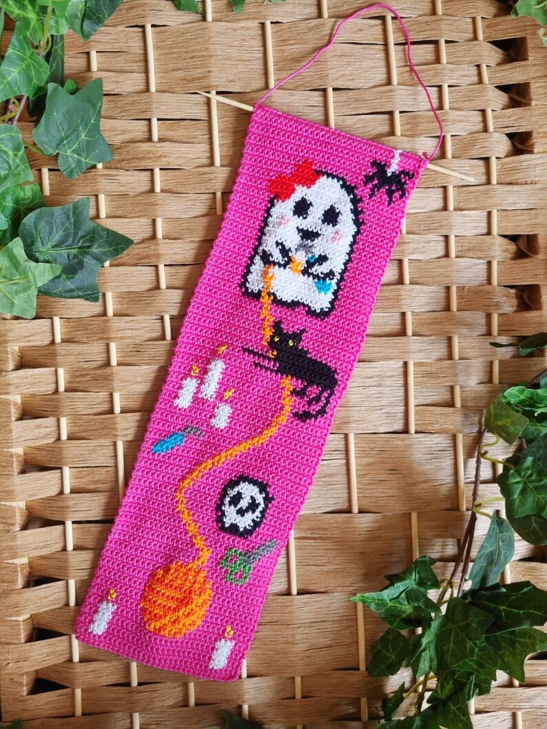 K&ouml;nnte beinhalten: Ein handgefertigter, pinkfarbener Lesezeichen zum Thema Halloween. Das Lesezeichen zeigt einen wei&szlig;en Geist mit roter Schleife, eine schwarze Katze, eine Spinne, Kerzen, einen Totenkopf und einen K&uuml;rbis. Das Lesezeichen ist aus H&auml;kelmaterial und hat eine pinkfarbene Aufh&auml;ngeschnur.