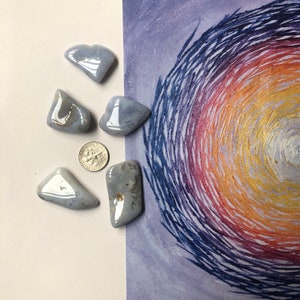 May include: Six polished, light blue gemstones with a glossy finish are arranged on a white surface next to a U.S. dime. The gemstones vary in shape and size. A colorful abstract painting with swirling brushstrokes in shades of purple, orange, and yellow is in the background.