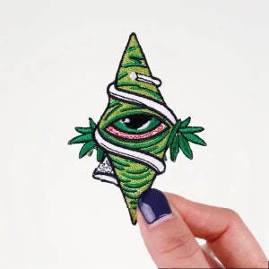 Peut inclure: Patch brodé vert avec un œil stylisé entouré d'un motif de feuille de marijuana. L'œil est vert avec une paupière rouge et une sclère blanche.