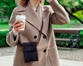 May include: A woman wearing a brown double-breasted coat with a black leather crossbody bag and sunglasses. She is holding a white coffee cup.