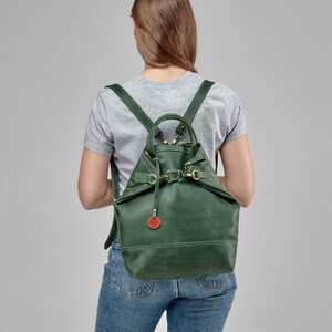 May include: A dark green leather backpack with adjustable straps and a buckle closure. The backpack has a small, round, red leather tag attached to it.