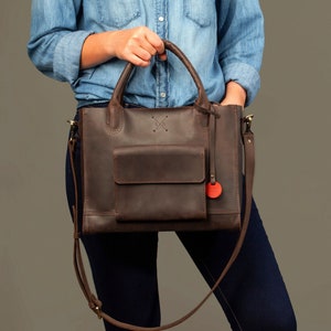 May include: A brown leather tote bag with a top handle and a long shoulder strap. The bag has a front pocket and a small red circle on the front.