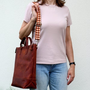 May include: A brown leather tote bag with a zipper closure and a white and orange checkered strap. The bag is being worn over the shoulder.