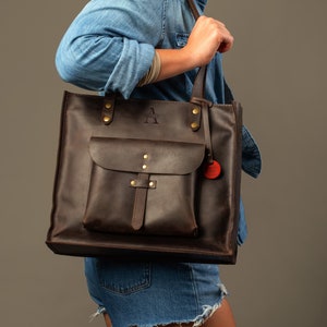 May include: A dark brown leather tote bag with a front pocket and a shoulder strap. The bag has brass hardware and a small red charm. The letter "A" is embossed on the upper part of the bag.