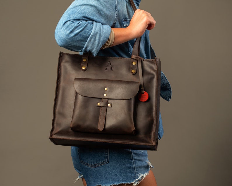 May include: A dark brown leather tote bag with a front pocket and a shoulder strap. The bag has brass hardware and a small red charm. The letter "A" is embossed on the upper part of the bag.