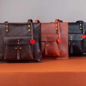 May include: Three leather tote bags in varying shades of brown and black. Each bag features a front pocket, top handles, and a decorative red tag. The bags are displayed on an orange surface against a gray backdrop.