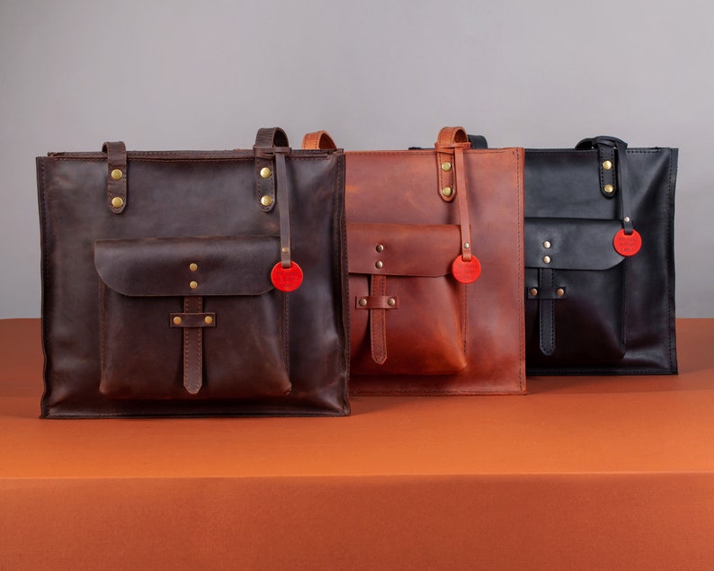 May include: Three leather tote bags in varying shades of brown and black. Each bag features a front pocket, top handles, and a decorative red tag. The bags are displayed on an orange surface against a gray backdrop.