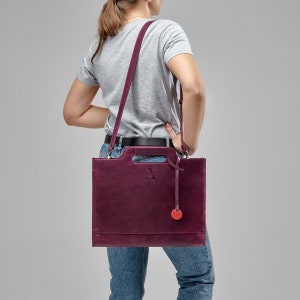 Puede incluir: Un bolso tote de cuero burdeos con asa superior y una larga correa para el hombro. El bolso tiene una peque&ntilde;a etiqueta circular roja unida a &eacute;l. El bolso tiene una letra "A" en relieve en la parte delantera.