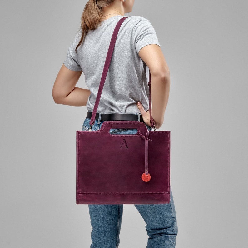 Puede incluir: Un bolso tote de cuero burdeos con asa superior y una larga correa para el hombro. El bolso tiene una peque&ntilde;a etiqueta circular roja unida a &eacute;l. El bolso tiene una letra "A" en relieve en la parte delantera.
