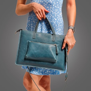 May include: A woman is holding a blue leather tote bag with a smaller pocket on the front. The bag has a long strap and is worn over the shoulder.