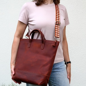 May include: A brown leather tote bag with a checkered orange and white strap. The bag has two top handles and a zipper closure.
