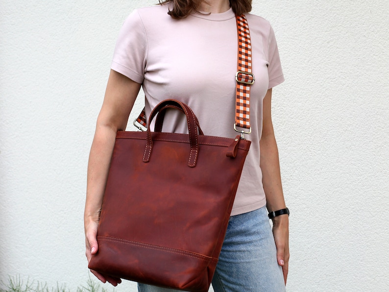 May include: A brown leather tote bag with a checkered orange and white strap. The bag has two top handles and a zipper closure.