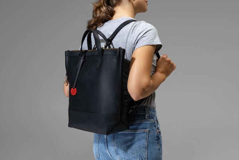 Op de afbeelding: Een zwarte leren tote bag met een rood rond label. De tas heeft een ritssluiting en verstelbare banden die als rugzak gedragen kunnen worden.