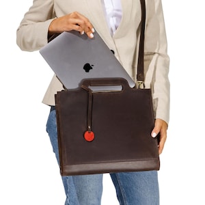 May include: A dark brown leather satchel with a top handle and a detachable shoulder strap. The bag has a zipper closure and a small red tag. A silver laptop is being placed inside the bag.