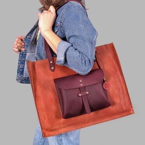 Op de afbeelding: Een bruine leren tote bag met een bordeauxrode leren zak en een klein bordeauxrood leren label met de tekst "The Maker's Goods".