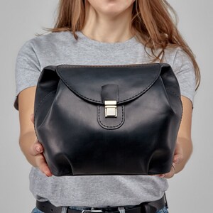 May include: A black leather handbag with a curved flap and a gold-colored clasp. The bag is held by a person, showcasing its shape and design. The stitching is visible, adding detail to the accessory.