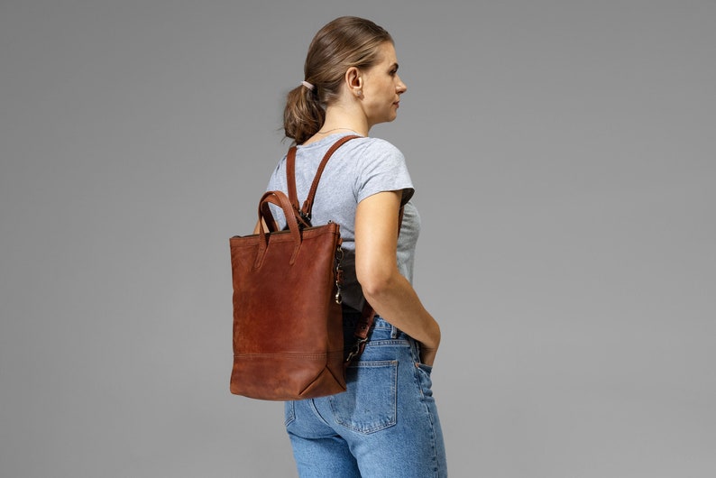 May include: A brown leather backpack with a tote bag style top. The bag has a zipper closure and a strap with a buckle. The backpack is being worn by a person wearing blue jeans and a gray t-shirt.
