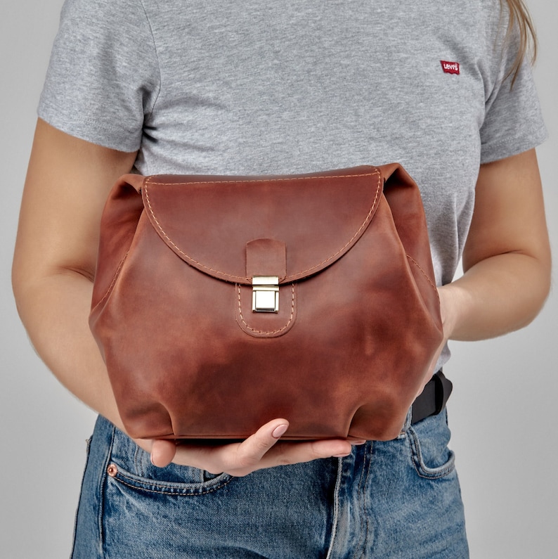 May include: A brown leather bag with a flap closure and a gold-colored clasp. The bag is held by a person wearing a gray t-shirt and blue jeans. The leather has a distressed look.