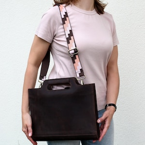 May include: A brown leather tote bag with a pink and white patterned strap. The bag has a top handle and a shoulder strap.