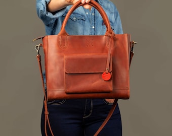 May include: A brown leather tote bag with a top handle and a long shoulder strap. The bag has a front pocket and a small red tag with the text "Handmade with Love" on it.