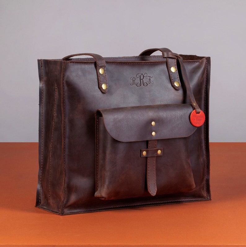 May include: A brown leather tote bag with a front pocket and top handles. The bag features gold-colored rivets and a small, red tag. The initials "RRF" are embossed on the front. The bag is designed for carrying items.