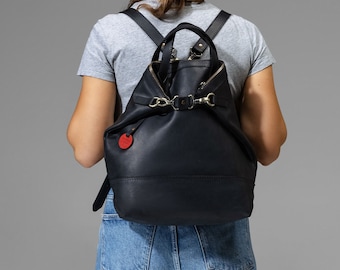 May include: A black leather backpack with a flap closure and silver hardware. The backpack has a red circle tag attached to it.