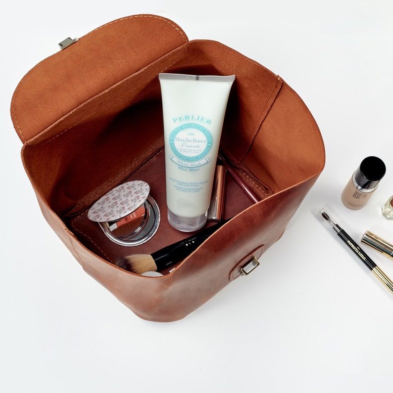 May include: A brown leather makeup bag, open to reveal various cosmetic products. Inside are a tube of white cream, a compact, a makeup brush, and lipsticks. Additional makeup items are arranged outside the bag on a white surface.