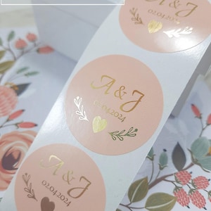 Puede incluir: Rollo de pegatinas redondas de color rosa pálido con letras y detalles dorados. Cada pegatina presenta las iniciales "A & J" y la fecha "02.04.2024", junto con un corazón y elementos florales. El texto "LITTLESTICKER CO" es visible arriba a la izquierda.