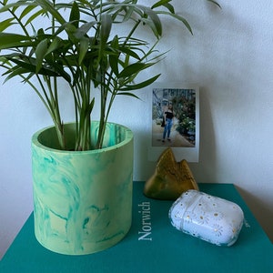 May include: A green and white marbled planter with a green leafy plant inside. The planter is sitting on a teal surface with a gold decorative object and a white phone case with a daisy pattern.