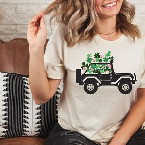 May include: A white t-shirt with a black silhouette of a jeep with green shamrocks on top. The jeep is driving on a road.