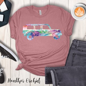 May include: A heather orchid t-shirt with a floral print of a jeep silhouette. The shirt is laid out on a wooden surface with a pair of black sneakers, a pair of jeans, and a cup of coffee.