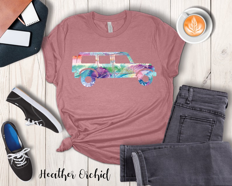 May include: A heather orchid t-shirt with a floral print of a jeep silhouette. The shirt is laid out on a wooden surface with a pair of black sneakers, a pair of jeans, and a cup of coffee.
