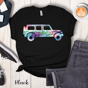 May include: Black t-shirt with a silhouette of a jeep with a tropical leaf pattern inside the silhouette. The jeep is facing right. The shirt is on a white wooden surface with a pair of black shoes, a brown leather notebook, a white phone, a cup of coffee, and a pair of gray jeans.