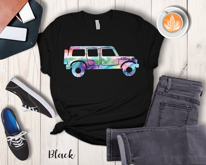 May include: Black t-shirt with a silhouette of a jeep with a tropical leaf pattern inside the silhouette. The jeep is facing right. The shirt is on a white wooden surface with a pair of black shoes, a brown leather notebook, a white phone, a cup of coffee, and a pair of gray jeans.