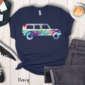 May include: A navy blue t-shirt with a colorful floral print of a jeep silhouette. The shirt is displayed on a white wooden surface with jeans and sneakers.