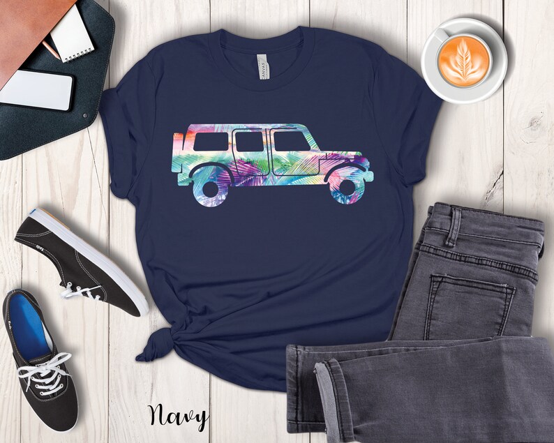 May include: A navy blue t-shirt with a colorful floral print of a jeep silhouette. The shirt is displayed on a white wooden surface with jeans and sneakers.