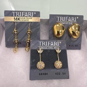 Könnte beinhalten: Eine Sammlung von Trifari-Ohrringen, die auf grauen Karten präsentiert werden. Die Ohrringe umfassen goldgefüllte Herz-Ohrhänger, goldene Komfort-Clip-Ohrringe und goldfarbene Ohrringe mit einem kristallbesetzten Kugeldesign. Die Karten zeigen das Trifari-Logo.