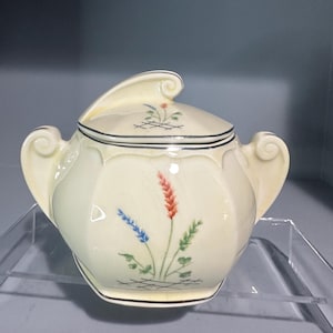 May include: Cream-colored ceramic sugar bowl with lid. The bowl features a floral design with red, blue, and green accents. It has two handles and a decorative lid with a curved handle. The rim is outlined with black.