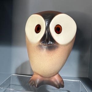 May include: A ceramic owl figurine with a brown and cream color scheme. The owl has large, round, cream-colored eyes with dark orange pupils. The owl's body is a light tan color, and the feet are brown.