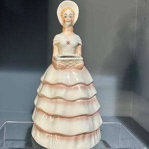 Może przedstawiać: Vintage ceramiczna figurka kobiety w białej sukni z różowymi wykończeniami i pasującym kapeluszem. Figurka trzyma kosz i ma szczegółową twarz. Suknia ma wiele warstw z falbanami. Mały różowy kwiat jest na piersi.