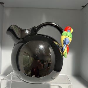 Puede incluir: Una jarra de cerámica negra brillante con cuerpo redondo y asa curva. El asa está adornada con una colorida figura de loro en rojo, amarillo, azul y verde. La jarra se exhibe sobre una estantería de acrílico transparente.