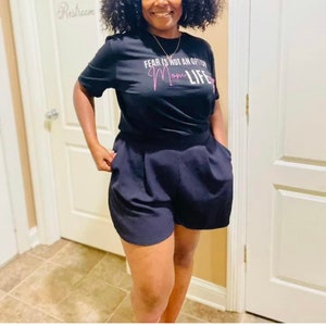 May include: A woman wearing a black t-shirt with the text "FEAR IS NOT AN OPTION Mom LIFE" in pink lettering. She is also wearing black shorts.