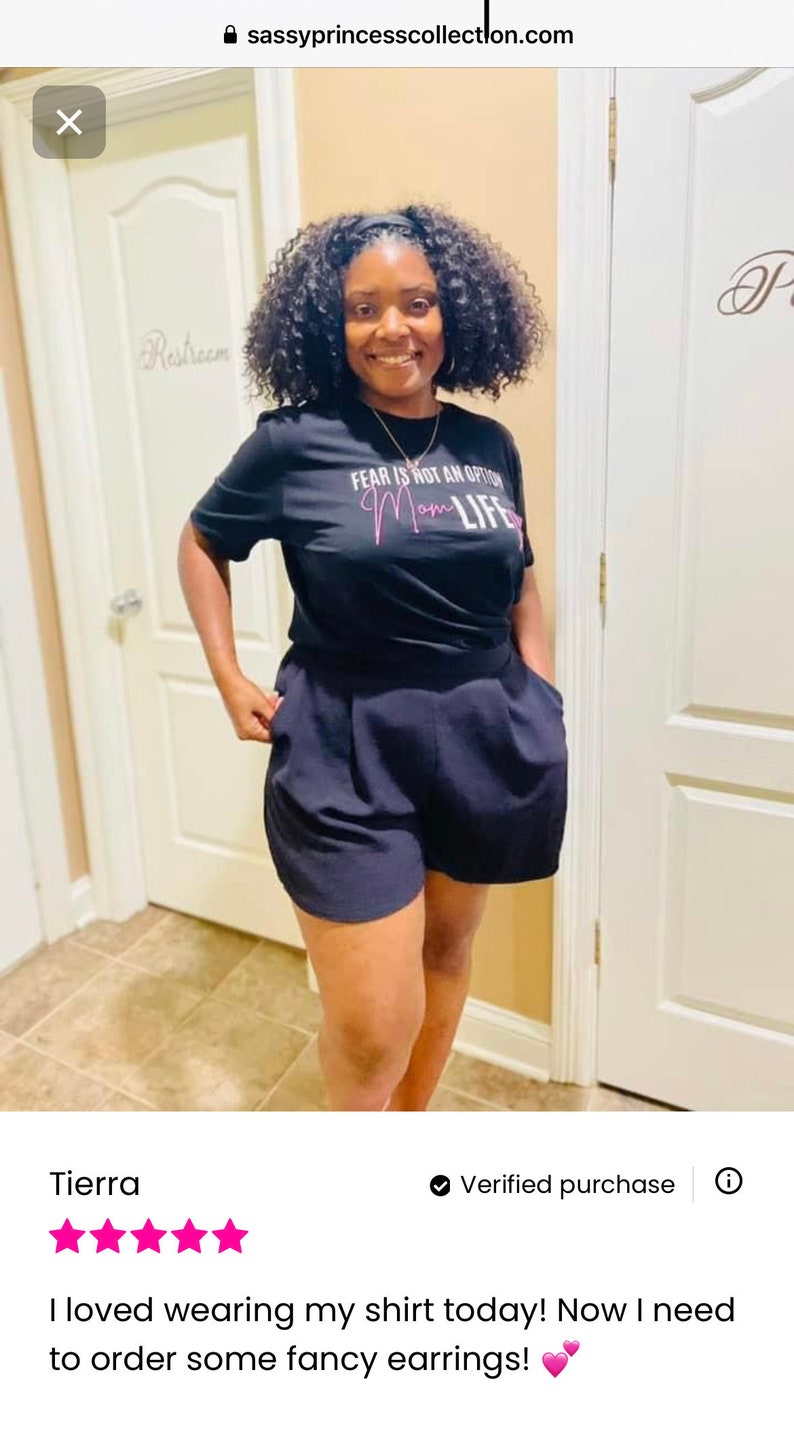 May include: A woman wearing a black t-shirt with the text "FEAR IS NOT AN OPTION Mom LIFE" in pink lettering. She is also wearing black shorts.