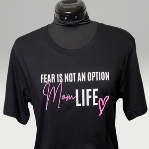 May include: Black t-shirt with white text that reads "Fear is not an option Mom Life" with a pink heart. The text "Mom Life" is in a pink script font.