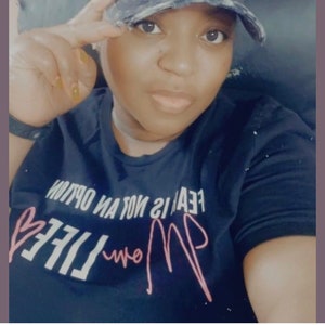 May include: A black t-shirt with the text "FEAR IS NOT AN OPTION... MOM LIFE" in pink lettering with a pink heart. The person wearing the shirt is also wearing a black and white baseball cap with the text "MOM LIFE" in pink lettering with a pink heart.